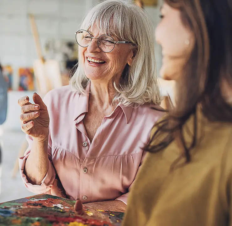 Three women enjoying painting together in a bright art studio; the focus is on a smiling older woman with glasses.