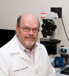 UCI coronavirus expert Michael Buchmeier dressed in a white lab coat sits in front of lab equipment and a laptop.