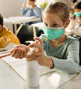 Sitting at a desk in a classroom, a young blonde girl in mask and green shirt squirts hand sanitizer into her hand.