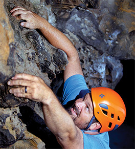 Brandon Gaidano dressed in a blue t-shirt and orange helmet grips the cliff face as he thinks about his next rock climbing move.