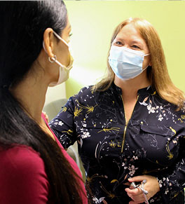 Dr. Lauri Seymour in a black, long-sleeved shirt with stethoscope in hand talks to brunette female patient in exam room. Both are masked.