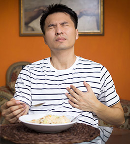 An Asian man sitting at the dinner table with a hand on his chest and a painful expression.