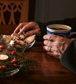 Hands of an elderly man sitting alone at a table with a mug, christmas cookies and candle.