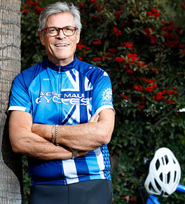 Michael Feldman, wearing a blue biking outfit, is leaning against a palm tree with his bicycle in the background.