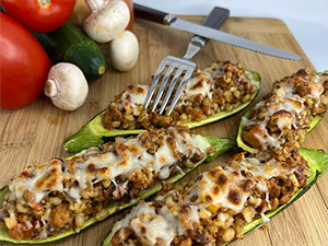 Four barley and sausage stuffed zucchini on a cutting board with fresh tomato and mushroom in the background.