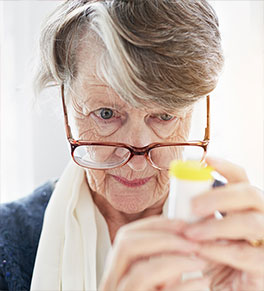 An older woman peers over her lowered glasses in an effort to read the label and instructions on a pill bottle.