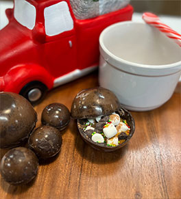An assortment of chocolate bombs on a cutting board with white mug, peppermint spoon and red toy truck.