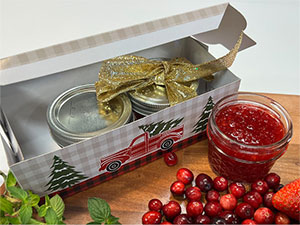 An open jar of Christmas jam next to a box of two other jars amidst fresh cranberries and mint.