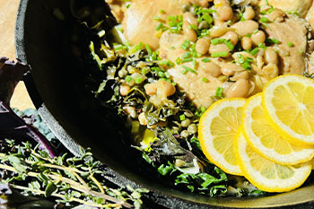 Braised chicken thighs in a black pan garnished with herbs and slices of lemon.