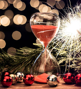 Red sand pours through an hourglass surrounded by ornaments, a sparkler and festive lights.