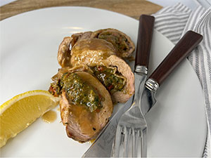 Slices of stuffed pork tenderloin covered in sauce displayed on a white plate with lemon garnish.