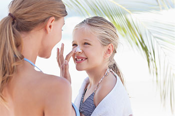 A blonde mother is spreading sunscreen on her daughter's noise at the beach.