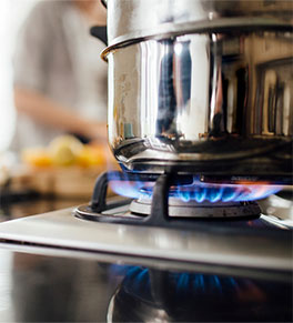 A pot is heating on a gas stove while a woman prepares food in the background.