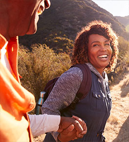A laughing Black woman is holding a man's hand to help him up a hill that they are hiking. 