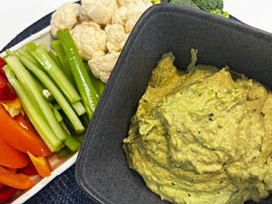 Green hummus is displayed in a gray square bowl with a side plate of red pepper, celery and cauliflower pieces.