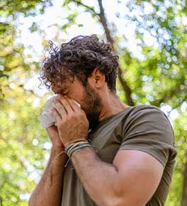 A young man sneezes into a tissue while hiking in a forest.