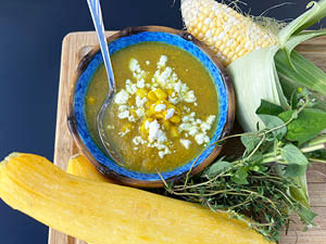 Golden Summer Corn Chowder served in a bright blue lowl surrounded by crookneck squash, corn on the cob and fresh herbs.