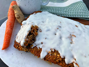 Oatmeal carrot cake with icing is served on white platter with a raw carrot, piece of ginger, speckled coffee mug and green checked towel in the background.
