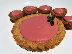 Raspberry curd tartlets with a golden almond crust displayed on a white platter on an outside table.