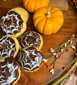 Spider web cookies are displayed with mini pumpkins on a wood cutting board.