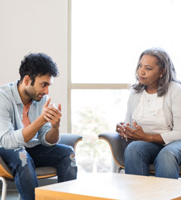 a young man and his therapist sit near a window at a table in chairs and discuss his depression