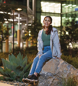 A night shot of smiling Kassandra Gomez sitting on big rock outside of a well-lit building.