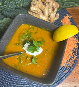 Red lentil soup garnished with yogurt and cilantro is in a gray stoneware bowl surrounded by raw kale, red lentils, ginger and turmeric.