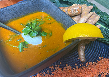 Red lentil soup garnished with yogurt and cilantro is in a gray stoneware bowl surrounded by raw kale, red lentils, ginger and turmeric.