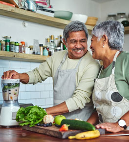 hispanic man and women in their 60s cooking a healthy meal full of vegetables in their home kitchen, laughing; man is starting the blender full of vegetables and fruits