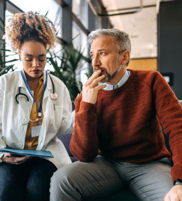 a man in a well lit room wearing red sweater and grey pants has just received a cancer diagnosis looking concerned outside sitting next to his female doctor an african american woman with a white coat and stethoscope
