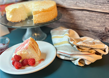 Angel Food Cake with Raspberry Drizzle is displayed on white plate with fresh berries on the side and the full cake in the background.