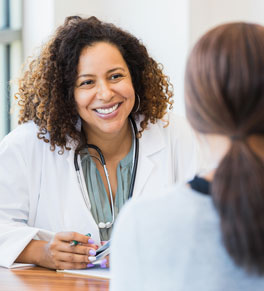 a female physician wearing a white coat, green shirt and stethoscope smiles at a female patient who has her hair in a ponytail