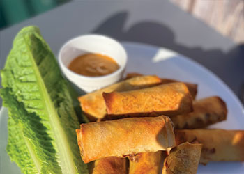 A batch of Balinese lumpia on a white plate with tamarind peanut dipping sauce and romaine lettuce on the side.