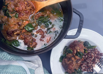 A large black pot of Jamaican oxtail stew with callaloo sits next to plated version served with wild rice.