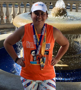 inflammatory bowel disease patient rocio castrillon with hands on hips wearing medals white hat black and white leggings and orange tank in front of a water fountain after running in a race