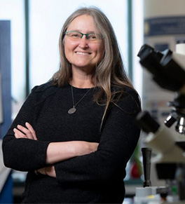 UCI scientist Katrine Whiteson, PhD, dressed in a black shirt, stands proudly in her lab with a big smile and arms crossed.