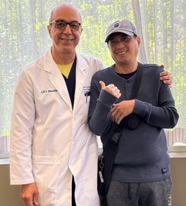 uci health medical oncologist dr arash rezazadeh in a white coat standing in front of a window with patient glenn marquez who is wearing a hat, dark sweater and dark pants while marquez smiles and points at rezazadeh