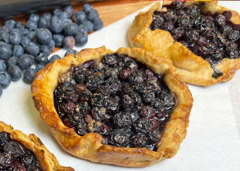 Mini blueberry tarts are displayed on white parchment paper with fresh blueberries in the background.