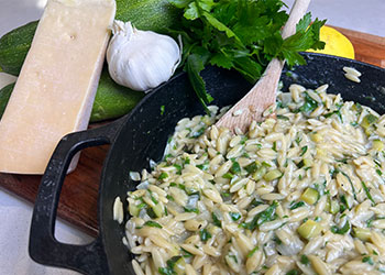 Lemony orzo with zucchini and parmesan in a cast iron skillet with fresh parmesan, zucchini, parsley, garlic and lemon in the background