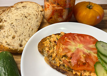 Open-faced kimchi salmon melt is served on white plate surrounded by cucumber, bread, jar of kimchi and whole tomato.