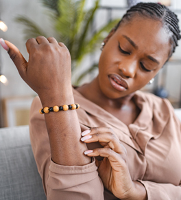 a woman sitting at home on her sofa wearing a bracelet and brown shirt scratching her right forearm