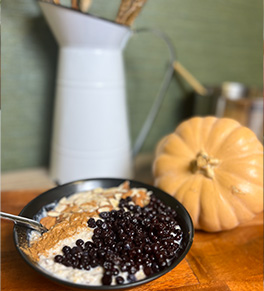 A creamy blueberry oatmeal bowl with almonds is served in a black bowl with a white pitcher and fresh mini-pumpkins in the background.