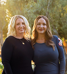 Sisters Rhea and Tara Hickman stand together smiling in golden sunset light.