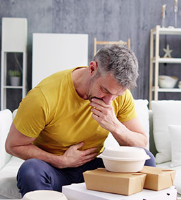 An older man sits on a sofa holding his stomach, hand on his mouth with boxes of fast food stacked on table in front of him.