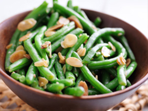 Roasted green beans with almond slivers is served in a brown bowl on a straw mat.