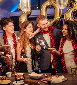 Two guys and two ladies in their 20s at holiday party lighting sparklers with appetizers and drinks on a table.