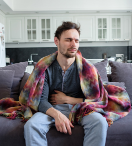 a man sitting on his sofa in front of the kitchen wearing a colorful blanket looking miserable holding his stomach