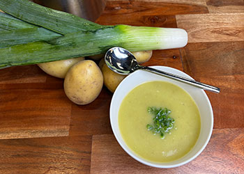 Leek and potato soup is displayed in a white bowl on a wood cutting board with fresh leeks and potatoes in the background.