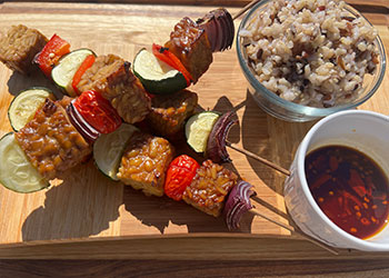 Grilled tempeh and veggie skewers are displayed next to bowls of wild rice and glaze.