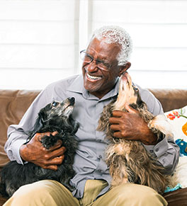 A laughing, older Black man is sitting on a leather couch with his two Dachshunds as one licks his ear.
